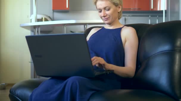 Красива молода жінка використовує ноутбук, сидячи в кріслі на кухні — стокове відео