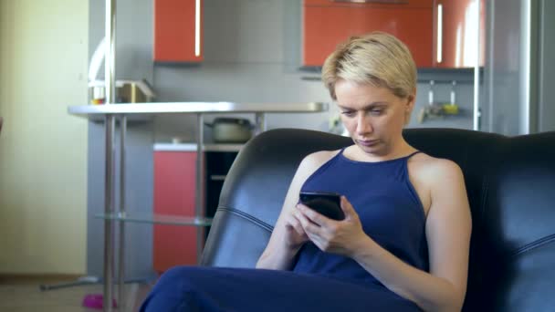 Verveelde vrouw gooit de telefoon zittend op een fauteuil in de woonkamer keuken — Stockvideo