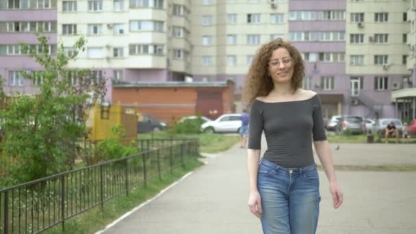 街の通りを歩く巻き毛のメガネの美しい笑顔の女性. — ストック動画