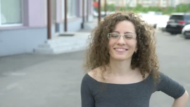 Bela mulher sorridente em óculos com cabelo encaracolado caminha em uma rua da cidade . — Vídeo de Stock