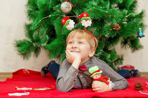 Щасливий усміхнений світло-волохатий маленький хлопчик лежить на червоній ковдрі під новорічною ялинкою з різдвяними прикрасами навколо нього — стокове фото