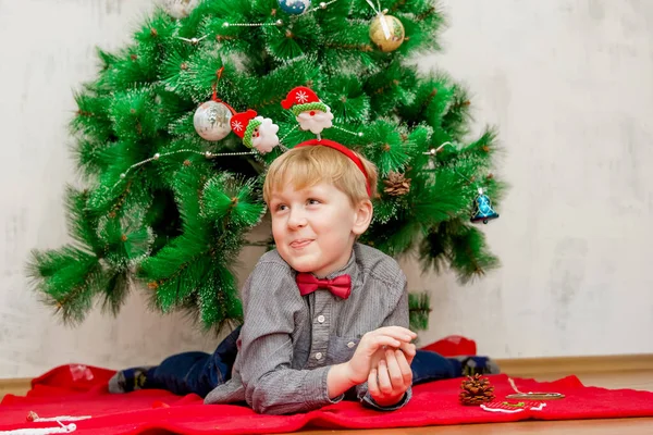 Щасливий усміхнений світло-волохатий маленький хлопчик лежить на червоній ковдрі під новорічною ялинкою з різдвяними прикрасами навколо нього — стокове фото
