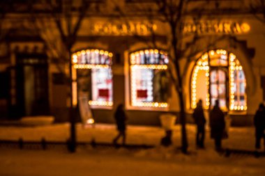 Şehir caddesinin noel ışıklarıyla çevrili pencerelerini göremiyoruz. Gece vakti. Kaldırımda yürüyen yayalar.