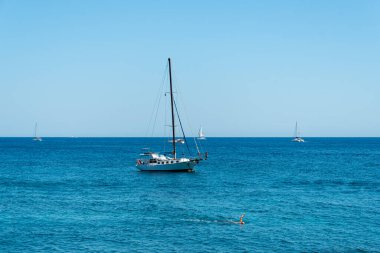 Akrotiri, Greece - July 19 2019:   A man swimming through the Aegean Sea waters passing a yacht off the seafront at Akrotiri
