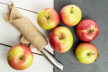 Gri taşın üzerinde altı tane taze bahçe elması ve beyaz ahşabın üzerinde kumaştan bir bıçak grubu.