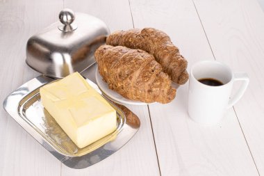 İki tam pişmiş kepekli kruvasan grubu ve beyaz ahşap üzerinde kahve ve tereyağı.