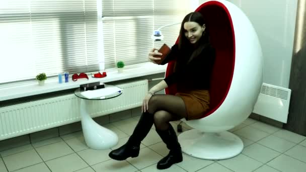 Uma jovem se senta em uma cadeira vermelha e tira uma selfie . — Vídeo de Stock