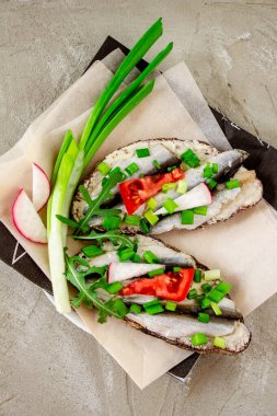 Çavdar ekmekli, tereyağlı, küçük sardalyeli, domates dilimli, turplu ve yeşil soğanlı lezzetli sandviçler. Beton parşömen üzerinde, sıra dışı bir yemek sunumu.