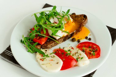 Bir dilim domates, mozzarella, roka, baharat ve beyaz bir tabakta siyah biber dilimlenmiş siyah çavdar ekmeğinin üzerinde nefis haşlanmış yumurta.