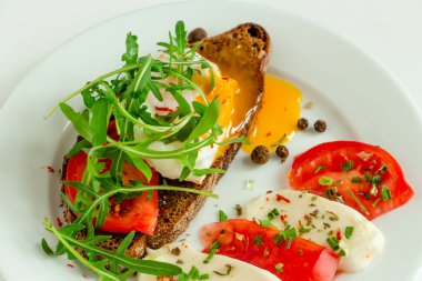 Bir dilim domates, mozzarella, roka, baharat ve beyaz bir tabakta siyah biber dilimlenmiş siyah çavdar ekmeğinin üzerinde nefis haşlanmış yumurta.