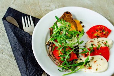 Lezzetli haşlanmış yumurta ve domates dilimleri, mozzarella, roka, baharat ve beton bir arka planda beyaz bir tabakta kara biber dilimleri.