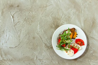 Lezzetli haşlanmış yumurta ve domates dilimleri, mozzarella, roka, baharat ve beton bir arka planda beyaz bir tabakta kara biber dilimleri.