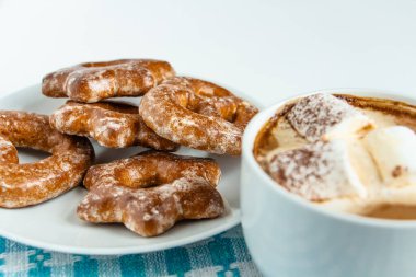 Beyaz bir tabakta kokulu zencefilli kurabiyeler bir fincan sıcak kakao ya da marşmelovlu kahve üzerine çikolata serpiştirilmiş, beyaz arka plan, mavi peçete, lezzetli tatlı, huzur hissi.