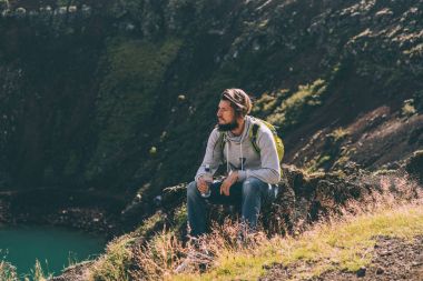 young bearded man sitting on rock and looking at beautiful crater lake in Iceland  clipart