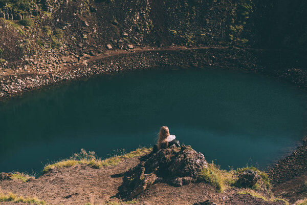 back view of young woman sitting on rock and looking at beautiful crater lake in Iceland 