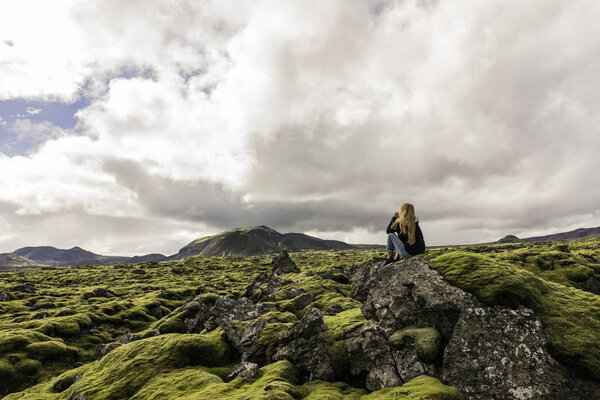 young woman sitting on rock and looking at majestic icelandic landscape