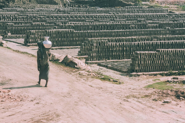 poor barefoot person carrying pitcher on shoulder and outdoor warehouse of bricks around in Nepal