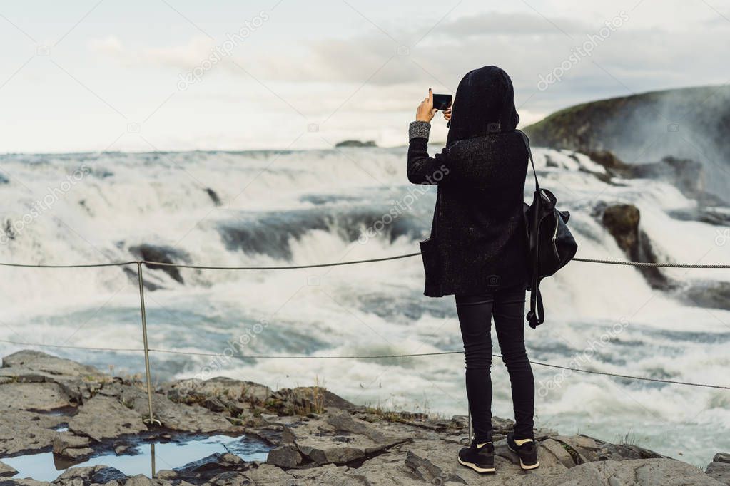 back view of young woman photographing majestic waterfall in Iceland 