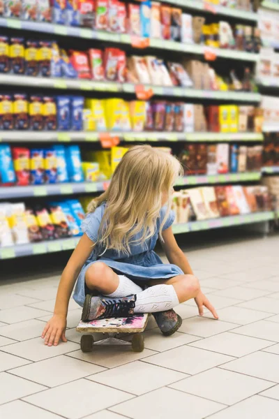 Anak Pirang Duduk Skateboard Supermarket Dengan Rak Belakang — Foto Stok Gratis