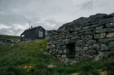 ruined old stone wall and rural house on background over field with green grass, Norway, Hardangervidda National Park clipart