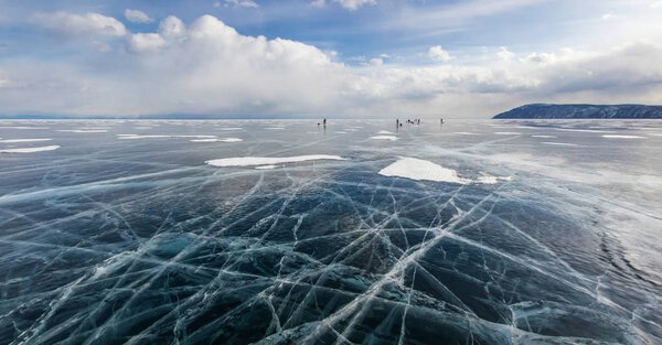 view of ice water surface under cloudy sky during daytime and group of hikers on background , russia, lake baikal  