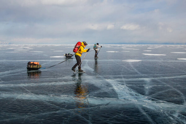 two men with backpacks going through ice water surface and hills on background, Russia, Lake Baikal