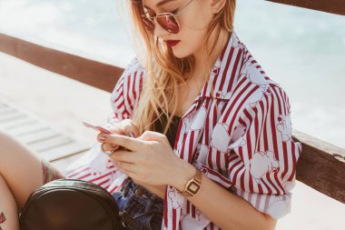 Smartphone açık havada pier bulanık deniz önünde katında otururken kullanan şık genç kadın
