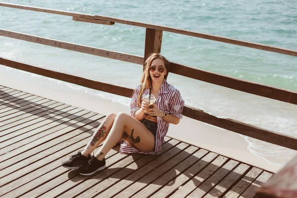 在海前的木码头上坐着塑料杯的笑的少妇 — 图库照片