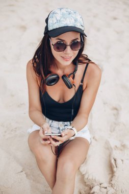 genç kadın kap ve akıllı telefon ve kulaklık rahatlatıcı kumsalda kullanarak güneş gözlüğü