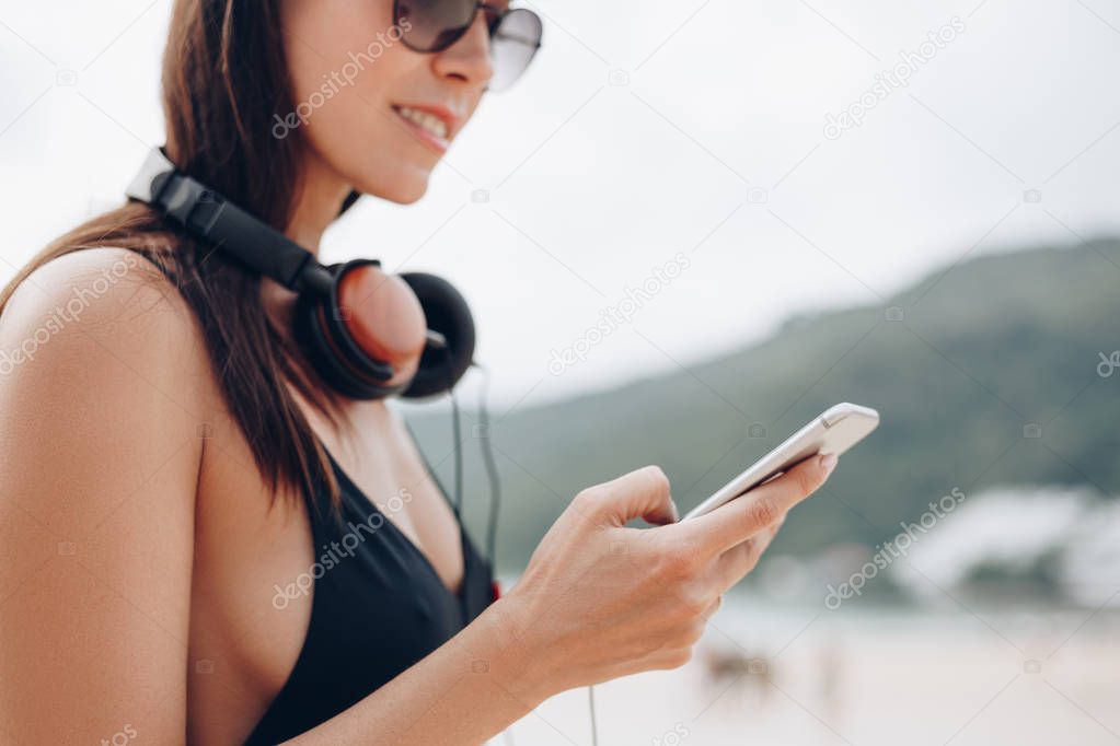 attractive woman in sunglasses and cap with headphones using smartphone