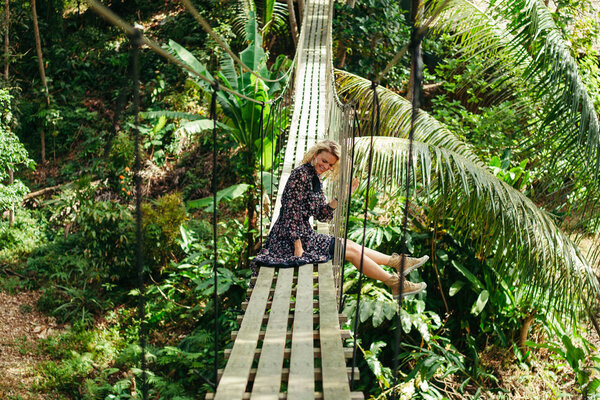 smiling attractive woman sitting on wooden footbridge in jungle