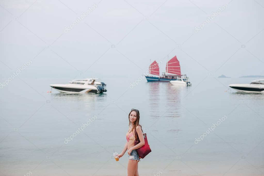 attraente ragazza con cocktail in piedi sulla spiaggia dell'oceano con yacht sullo sfondo