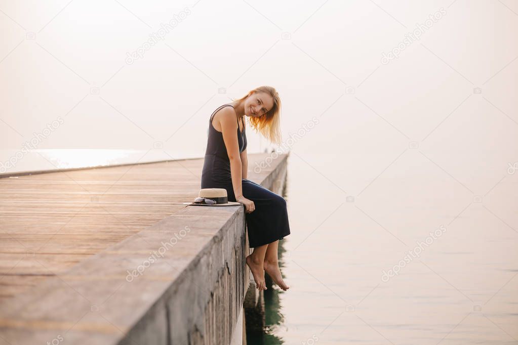 side view of smiling attractive girl sitting in dress on pier near ocean