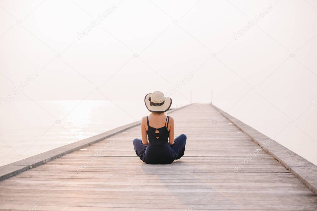 back view of woman in hat sitting on pier near ocean