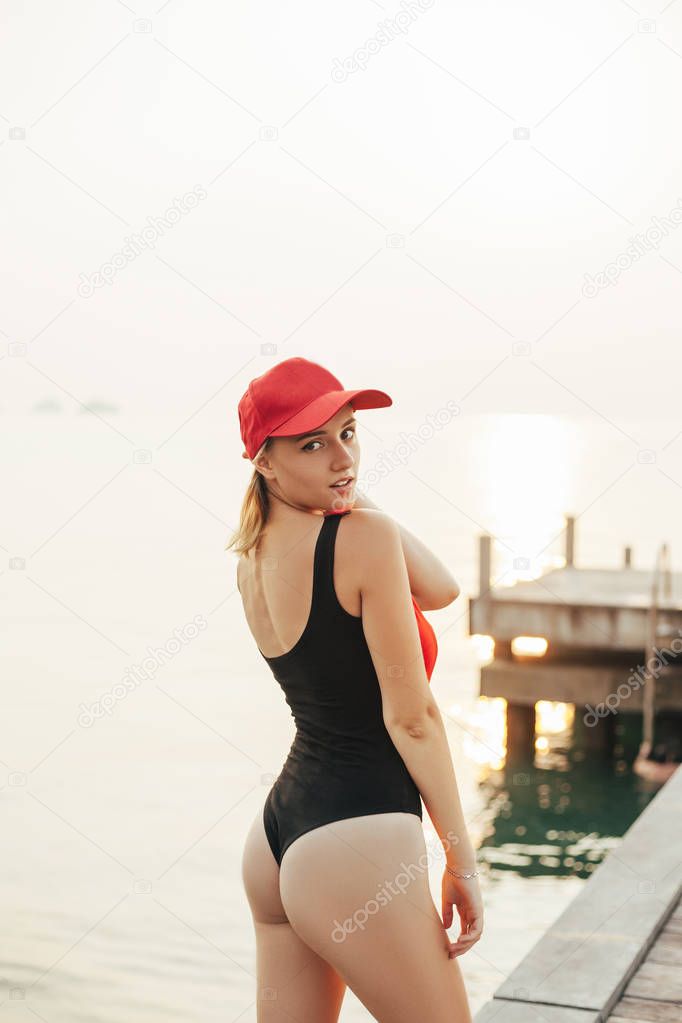 seductive beautiful girl posing in swimsuit and cap near ocean and looking at camera