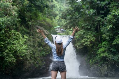 back view of woman with outstretched arms with Aling-Aling Waterfall on background, Bali, Indonesia clipart