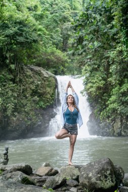 attractive woman practicing yoga with Aling-Aling waterfall on backdrop, Bali, Indonesia clipart