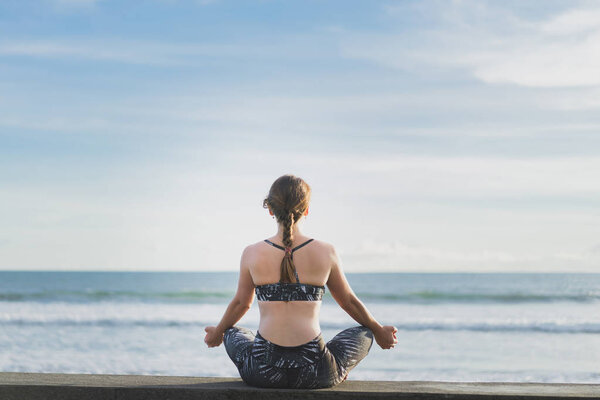 back view of woman meditating in lotus position with ocean and blue sky on background, Bali, Indonesia
