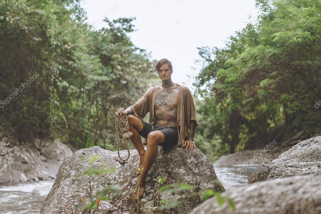 tattooed man with water bottle resting on rock with green plants and river on backdrop, Bali, Indonesia