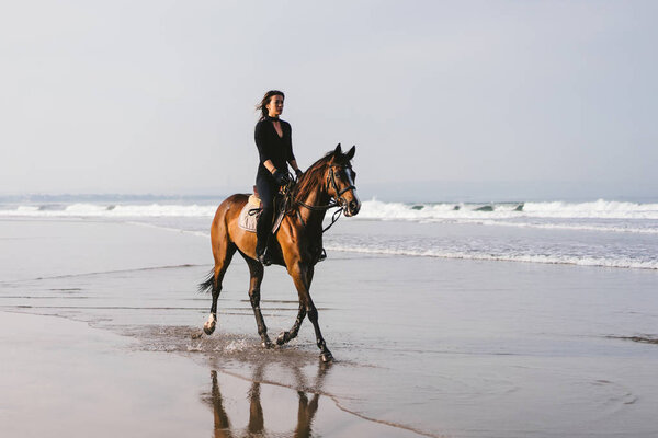 young female equestrian riding horse near wavy ocean