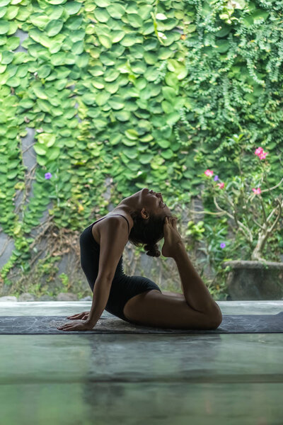 Side view of beautiful young woman practicing yoga and performing back bend with feet  touching head in front of wall covered with green leaves