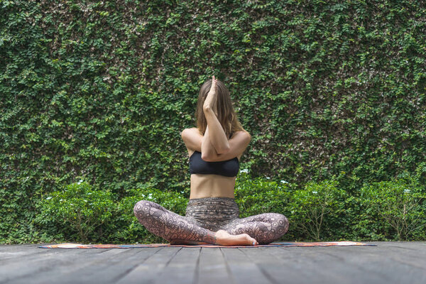 attractive young woman practicing yoga in lotus pose in front of wall covered with green leaves