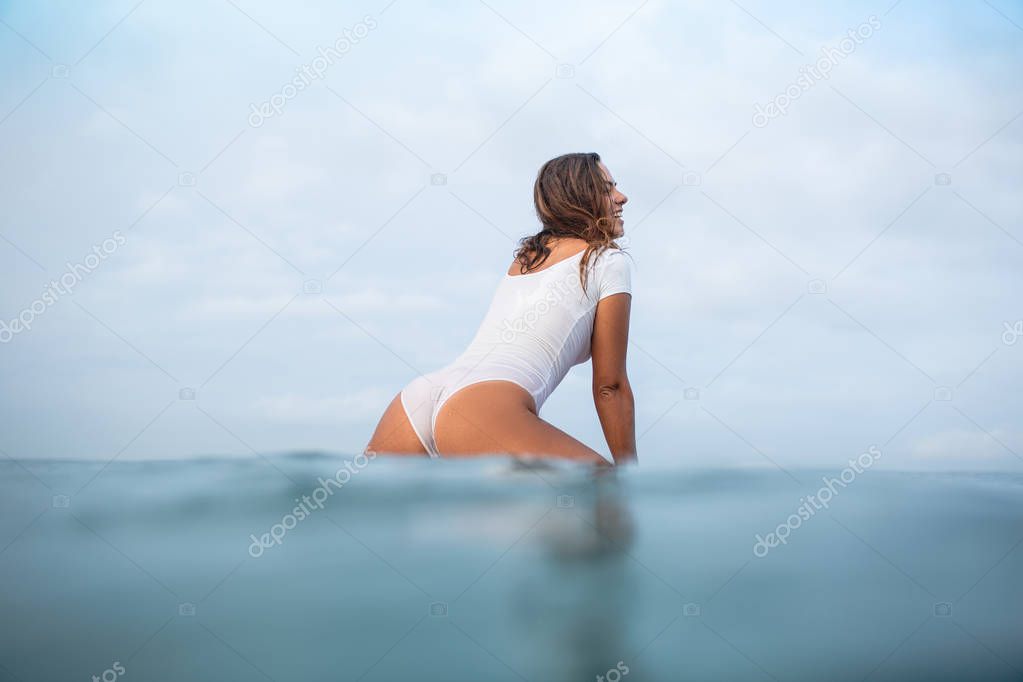 attractive young woman in wet white swimsuit sitting on surfboard in ocean