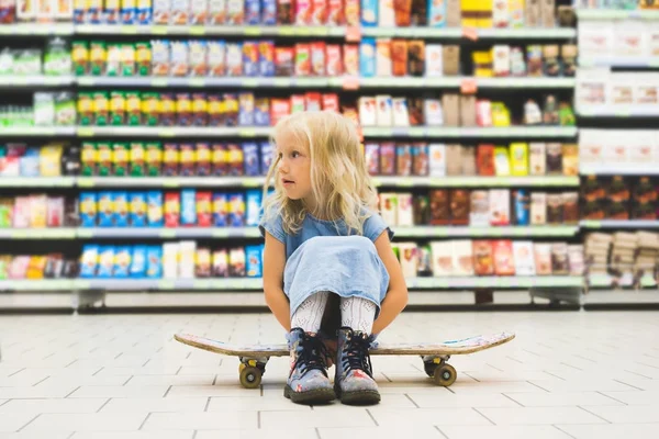 Little blonde child sitting on skateboard in supermarket with shelves behind — Stock Photo