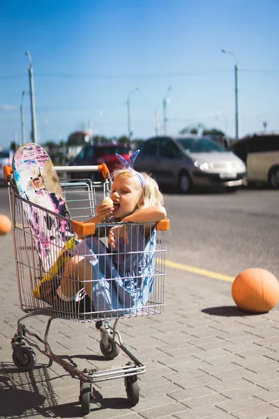 Pequeña niña comiendo patatas fritas mientras está sentada en el carrito de la compra con monopatín - foto de stock