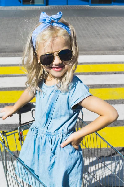 Little adorable female kid in sunglasses having fun in shopping cart on crosswalk — Stock Photo