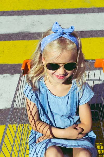 Criança elegante em óculos de sol sentado no carrinho de compras na passarela — Fotografia de Stock