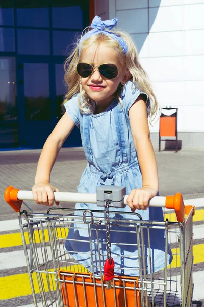 Little stylish child in sunglasses having fun in shopping cart at parking — Stock Photo