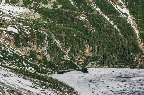 Vista de penhasco de montanha com árvores em encostas e lago a pé com gelo na superfície da água, Morskie Oko, Sea Eye, Tatra National Park, Polônia — Fotografia de Stock