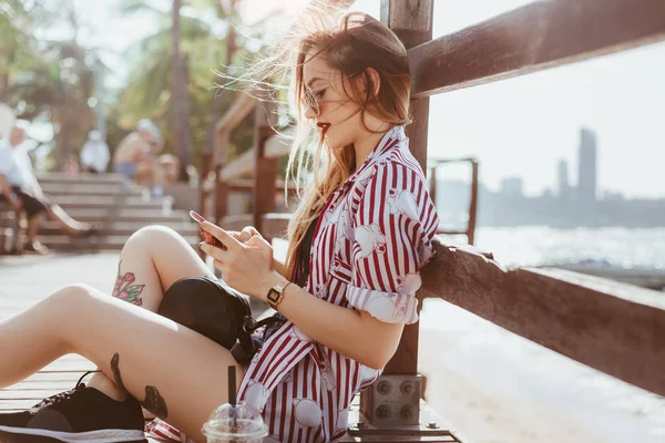 Beautiful young woman using smartphone while sitting on pier at beach — Stock Photo